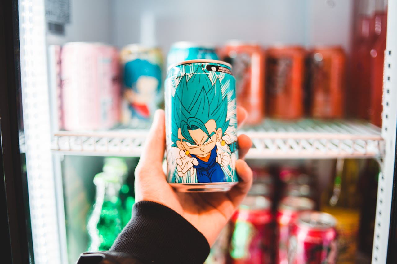 Close-up of a hand holding an anime character drink can in front of a cooler shelf.